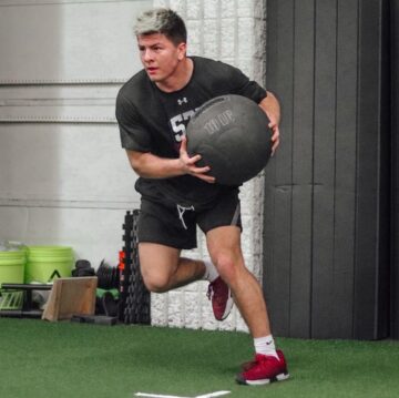An male athlete doing side-to-side one-legged jumps while holding a medicine ball.