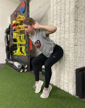 A female athlete doing a GOATA Bow Wall Sit Hold.