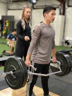 A coach helps a youth athlete with his form and shoulder position at the top of a trap-bar squat.