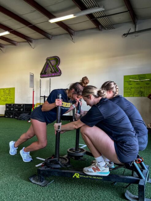 Two girls squatting on exercise sled, being pushed by another girl athlete.