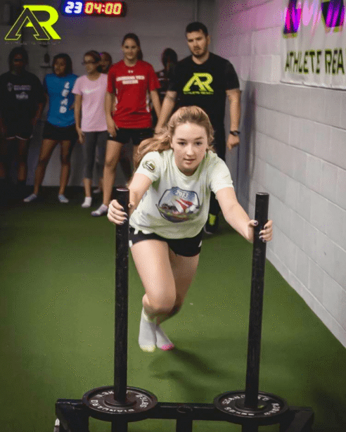 A female youth athlete pushing a sled in a gym with a group of other athletes and a coach watching her.